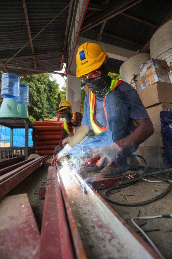 Construction worker welding metal beams outdoors with safety gear and hard hat.